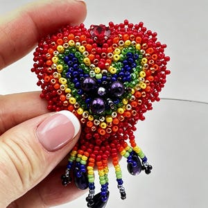 Op de afbeelding: Handgemaakte, hartvormige broche van kralen in levendige kleuren. De broche heeft een regenboogontwerp met rode, oranje, gele, groene, blauwe en paarse kralen. Het heeft een rode edelsteen aan de bovenkant en bungelende paarse kralen aan de onderkant.