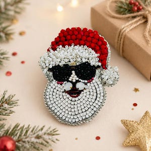May include: A festive Santa Claus brooch, crafted with beads. The Santa's hat is red, his beard is white, and he wears black sunglasses. The image includes Christmas decorations, such as a gift box and a gold star.
