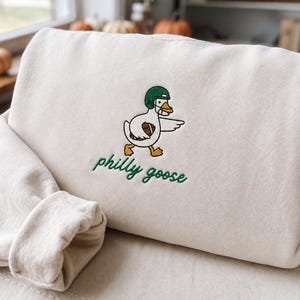 May include: A cream-coloured sweatshirt featuring an embroidered graphic of a white goose wearing a green football helmet and holding a brown football under its wing. Below the goose, the text philly goose is embroidered in green cursive script.