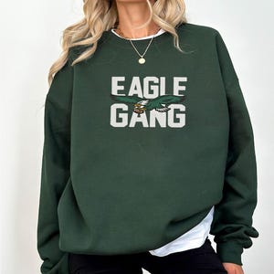 May include: A dark forest green crewneck sweatshirt featuring the white block text EAGLE GANG centred on the chest. Between the two words is a graphic illustration of a flying eagle with green and white feathers clutching a brown football.
