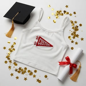 Könnte beinhalten: Ein weißes Crop-Top mit Spaghettiträgern und einem roten Wimpel mit der Aufschrift "CUSTOM". Eine schwarze Abschlusskappe mit goldener Quaste, ein gerolltes Diplom mit rotem Band und goldene Konfetti sind verstreut.