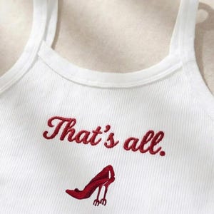 May include: White ribbed tank top with the embroidered phrase "That's all." and a red high-heeled shoe design. The top features a scoop neckline and thin straps, perfect for warm weather or layering.