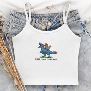 May include: White camisole top with thin straps. The front features a cartoon illustration of a blue dinosaur with a brown animal wearing a crown, with the text "THIS IS AN OUTRAGE" below the graphic. The top is laying on a pair of blue jeans.