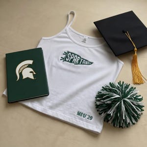 Pode incluir: Uma regata branca com a palavra "SPARTANS" em verde, um pompom verde e branco, um caderno verde com o logotipo de um capacete espartano e um chapéu de formatura preto com uma borla dourada. O texto "MSU '29" está na regata.