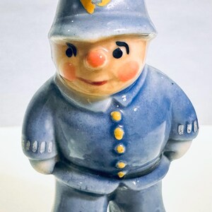 May include: A ceramic police officer figurine in a blue uniform and helmet. The figure has a rosy face, black eyes, and a small yellow badge on the helmet. The uniform features yellow buttons.