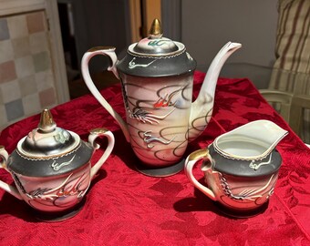 Op de afbeelding: Een vintage porseleinen theeservies met een theepot, suikerpot en melkkan. De set heeft een zwarte en gouden rand, met een roze en crème body. De theepot en suikerpot hebben gouden knoppen. De set is versierd met een drakenmotief.
