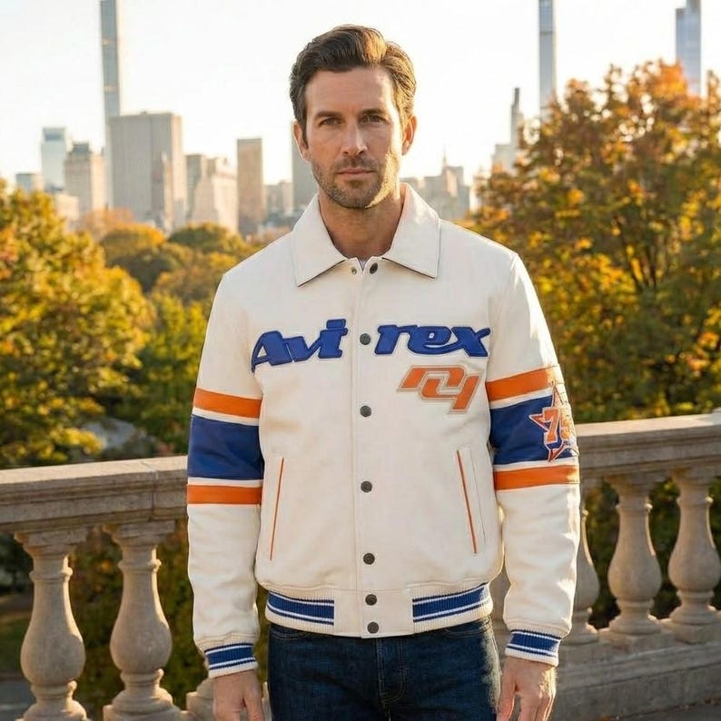 May include: Cream-colored jacket with blue and orange accents. The jacket features the brand name "Avi-rex" in blue lettering across the chest. The sleeves have blue and orange stripes, and the cuffs and hem are ribbed with blue and white stripes. The jacket is worn over blue jeans.