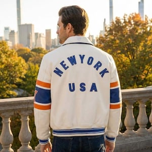 May include: Cream-colored jacket with "NEW YORK USA" lettering on the back. The jacket features blue and orange stripes on the sleeves and a blue and white striped hem. The jacket is worn outdoors with a city skyline in the background.