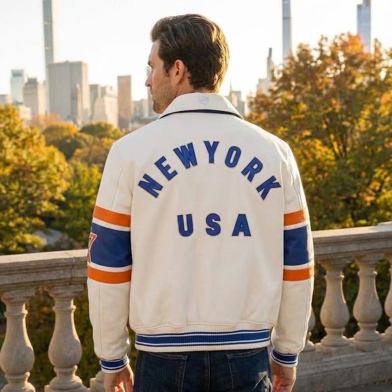May include: Cream-colored jacket with "NEW YORK USA" lettering on the back. The jacket features blue and orange stripes on the sleeves and a blue and white striped hem. The jacket is worn outdoors with a city skyline in the background.