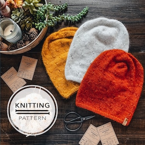 Pode incluir: Três chapéus de tricô em cores diferentes: amarelo mostarda, branco e laranja. Os chapéus estão dispostos sobre uma superfície de madeira com uma etiqueta que diz "Feito à mão com amor".