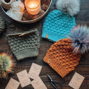 Puede incluir: Gorros de punto hechos a mano en color turquesa y naranja con pompones de piel sintética. Un gorro de punto verde en proceso y una colección de hilo y materiales de punto están dispuestos sobre una superficie de madera. También se ven etiquetas con instrucciones de cuidado.