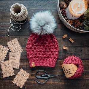 May include: A red knitted beanie with a white pom pom. The beanie has a textured stitch pattern and is handmade. The beanie is on a wooden surface with several small tags and a ball of yarn.
