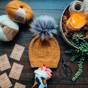 May include: A mustard yellow knitted beanie with a fluffy blue and purple pom pom. The beanie has a textured knit pattern and is handmade. The beanie is sitting on a wooden surface with yarn balls, a candle, and a pair of scissors.