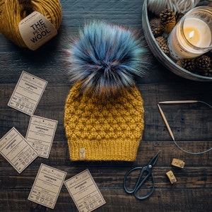 May include: A mustard yellow knitted beanie with a blue and purple faux fur pom pom. The beanie has a textured stitch pattern and a brown leather tag with the text "HANDMADE with love" and care instructions. The beanie is on a wooden surface with yarn, scissors, and other craft supplies.