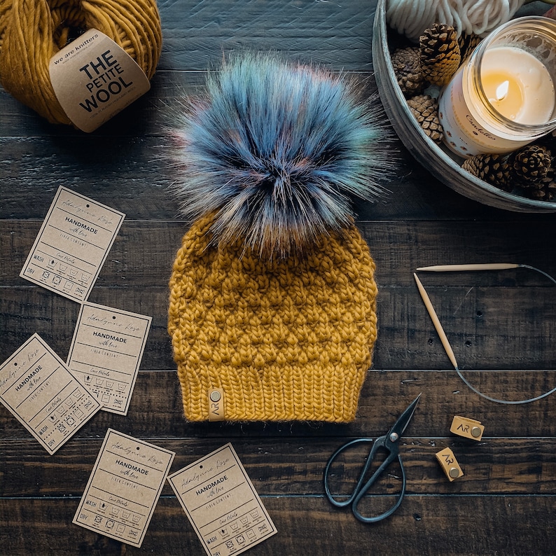 May include: A mustard yellow knitted beanie with a blue and purple faux fur pom pom. The beanie has a textured stitch pattern and a brown leather tag with the text "HANDMADE with love" and care instructions. The beanie is on a wooden surface with yarn, scissors, and other craft supplies.
