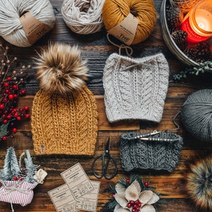 May include: Two knitted hats, one mustard yellow and one light grey, with a pom pom on the yellow hat. There are balls of yarn in the background, along with knitting needles and scissors. The image also includes care instructions for the hats.