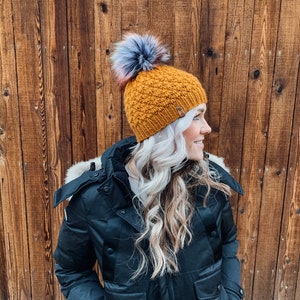 May include: A woman wearing a black winter coat with a fur-trimmed hood and a mustard yellow knit beanie with a multi-coloured pom-pom.