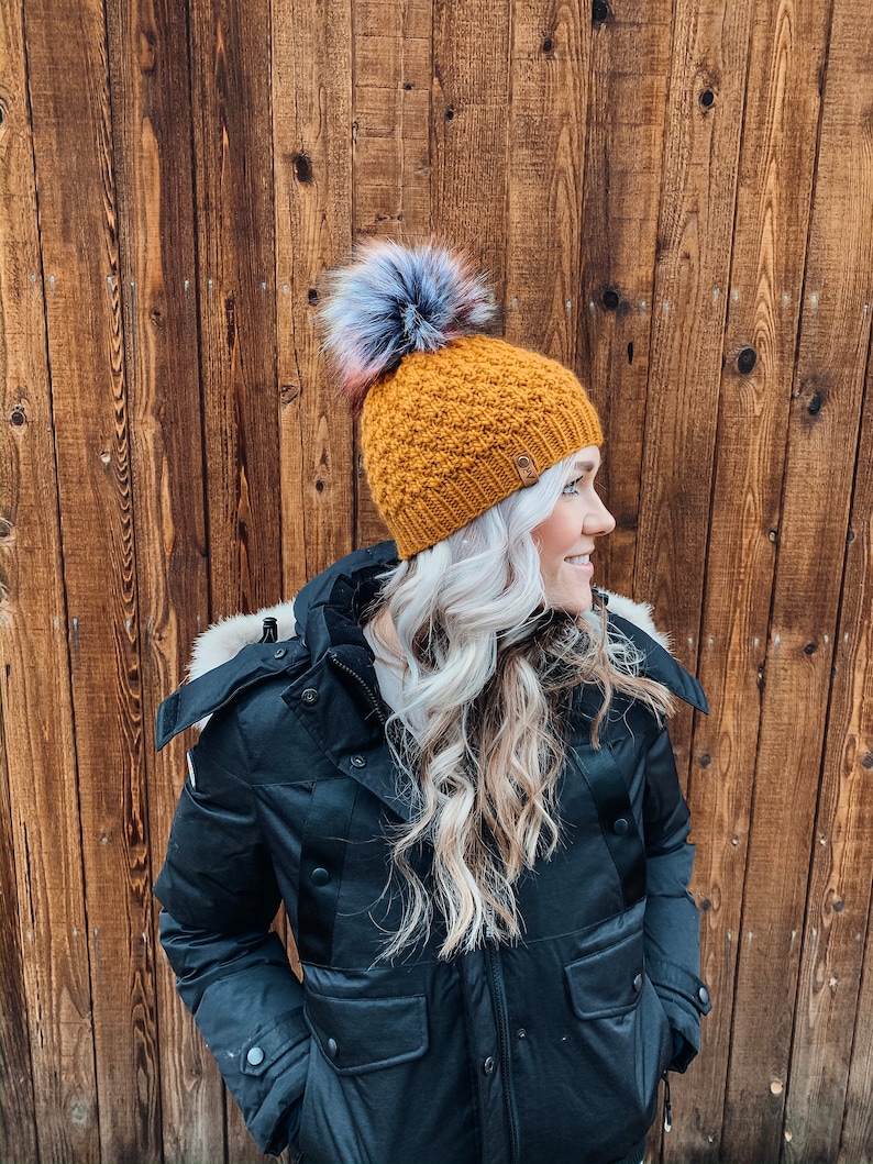 May include: A woman wearing a black winter coat with a fur-trimmed hood and a mustard yellow knit beanie with a multi-coloured pom-pom.