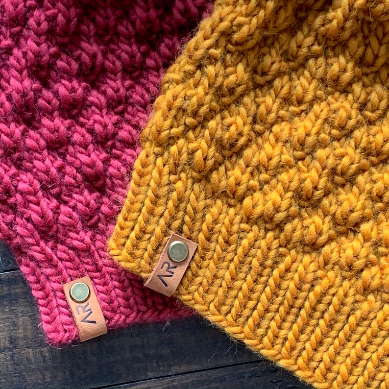 May include: Two knitted hats, one in a deep red colour and the other in a mustard yellow colour. Both hats have leather labels with the initials "AR" on them.