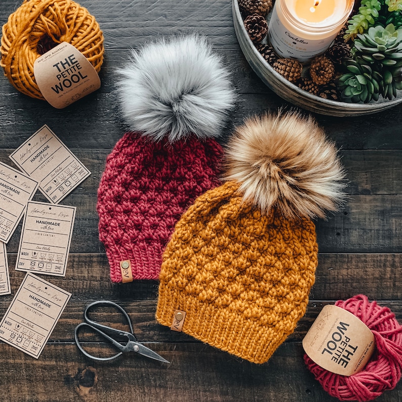 May include: Two handmade crocheted hats with pom poms. One hat is red and the other is mustard yellow. Both hats have care instructions tags attached.