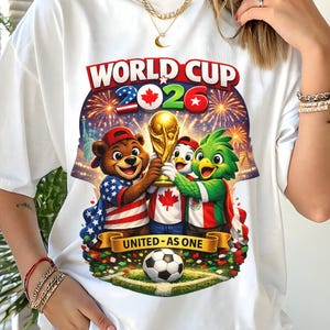 May include: White t-shirt featuring a colorful graphic for the 2026 World Cup. The design includes a bear, a penguin, and a parrot holding the World Cup trophy, with fireworks and the text "UNITED - AS ONE". A soccer ball is at the bottom.