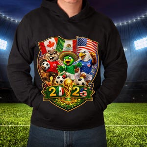 May include: A black hooded sweatshirt featuring a graphic print of a beaver, a parrot in a sombrero, and an eagle playing soccer. The design includes the flags of Canada, Mexico, and the USA, a gold trophy, and the text 2026 with flag accents.