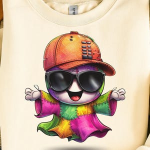 May include: A cream-colored sweatshirt featuring a colorful cartoon character wearing sunglasses and a baseball cap. The character is made of patchwork fabric in various colors, including pink, green, yellow, and purple. The character has its arms outstretched.