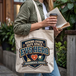 May include: A beige canvas tote bag featuring a graphic design with a shield shape, the text BEHIND EVERY SAFE SPACE EVS HERO ENVIRONMENTAL SERVICES, and icons of a broom, a heart, and a bucket with a cloth, printed in navy blue, teal, and orange.