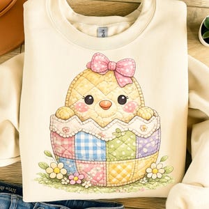 Può includere: Felpa color crema con un affascinante pulcino trapuntato che esce da un uovo patchwork. Il pulcino ha un fiocco rosa e guance rosee. Il guscio dell'uovo è decorato con quadrati pastello e accenti floreali, perfetto per Pasqua.