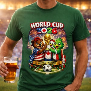 May include: Green t-shirt with a colorful graphic celebrating the 2026 World Cup. The design features cartoon mascots holding the trophy, a soccer ball, and the text "UNITED - AS ONE". The shirt is worn by a person holding a glass of beer.