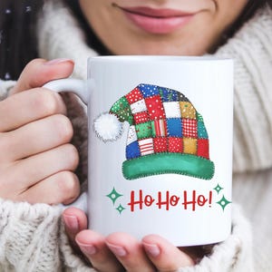 May include: White ceramic mug featuring a colorful patchwork Santa hat design with a white pom-pom and green trim. The mug has the text "Ho Ho Ho!" in red with green star accents, perfect for the holiday season.