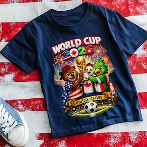 May include: Navy blue t-shirt with a colorful graphic featuring the text "WORLD CUP 2026" and three cartoon mascots holding a trophy. The mascots represent the USA, Canada, and Mexico. A soccer ball is also visible.