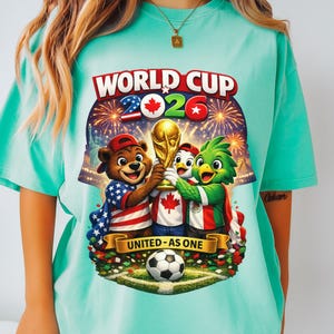 May include: A light teal t-shirt featuring a colorful graphic for the 2026 World Cup. The design includes cartoon mascots holding the World Cup trophy, fireworks, and the text "WORLD CUP 2026" and "UNITED - AS ONE". A soccer ball is at the bottom.