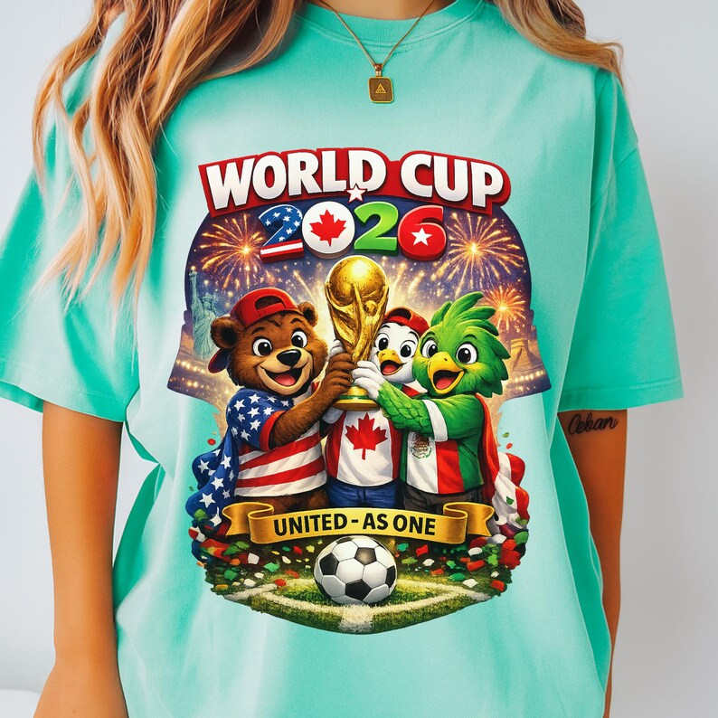 May include: A light teal t-shirt featuring a colorful graphic for the 2026 World Cup. The design includes cartoon mascots holding the World Cup trophy, fireworks, and the text "WORLD CUP 2026" and "UNITED - AS ONE". A soccer ball is at the bottom.