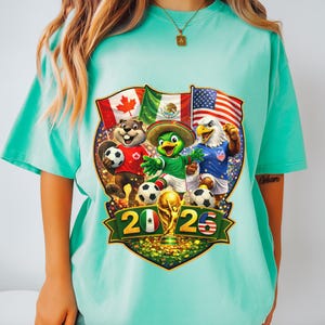 May include: A mint green t-shirt featuring a graphic of a beaver, a parrot, and an eagle playing soccer in front of the Canadian, Mexican, and American flags. The design includes the text 2026 and a gold trophy, with soccer balls at their feet.