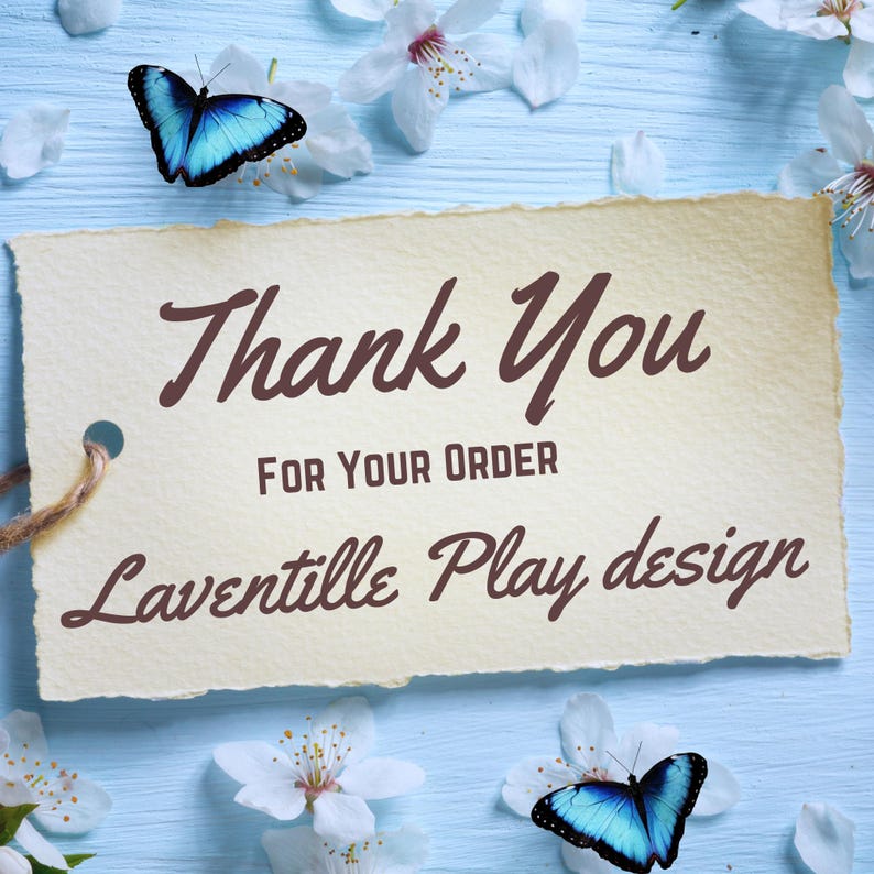 May include: A beige card with the words "Thank You FOR YOUR ORDER Laventille Play design" in brown script. The card is surrounded by white flowers, blue butterflies, and rests on a light blue wooden surface.