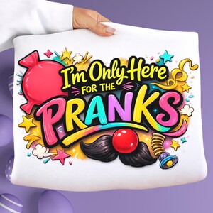 May include: A white sweatshirt featuring a colorful graphic design with the text I'm Only Here FOR THE PRANKS. The illustration includes a large pink candy, yellow stars, a black mustache, a red clown nose, and a spring-loaded toy element.