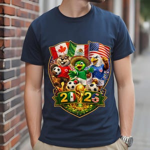 May include: A navy blue t-shirt featuring a graphic of a beaver, a parrot in a sombrero, and an eagle playing soccer. Behind them are the flags of Canada, Mexico, and the USA. Below, the text 2026 is displayed with a gold trophy in the center.