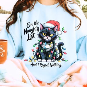 May include: Light blue sweatshirt featuring a black cat wearing a Santa hat and wrapped in Christmas lights. Text reads "On the Naughty List And I Regret Nothing."