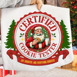 May include: White fabric featuring a circular design with Santa Claus holding a cookie and a glass of milk. The design reads "CERTIFIED COOKIE TESTER" in red and white, with the text "JOY, LAUGHTER, AND EVERYTHING BRIGHTER" on a red banner. Green Christmas trees frame the design.