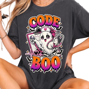 Könnte beinhalten: Ein anthrazitfarbenes T-Shirt mit einer Grafik eines als Krankenschwester verkleideten Geistes mit Stethoskop und Klemmbrett. Der Text CODE BOO ist in orange-violetten Verlaufsfarben gedruckt. Das Design zeigt kleine Fledermäuse, Sterne und ein EKG.