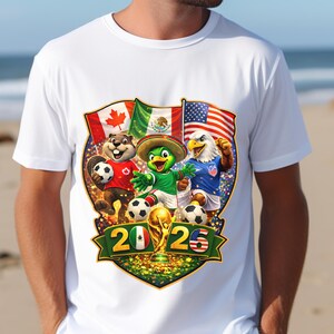 May include: A white t-shirt featuring a graphic of a beaver, a parrot, and an eagle playing soccer. Behind them are the flags of Canada, Mexico, and the USA. Below, the text 2026 is displayed with a gold trophy in the center on a green banner.