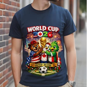 May include: Navy blue t-shirt featuring a colorful graphic for the 2026 World Cup. The design includes cartoon mascots holding the trophy, fireworks, and the text "WORLD CUP 2026" and "UNITED - AS ONE". A soccer ball is at the bottom.