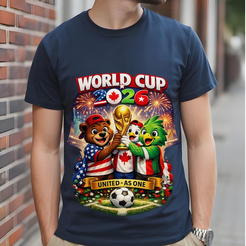 May include: Navy blue t-shirt featuring a colorful graphic for the 2026 World Cup. The design includes cartoon mascots holding the trophy, fireworks, and the text "WORLD CUP 2026" and "UNITED - AS ONE". A soccer ball is at the bottom.