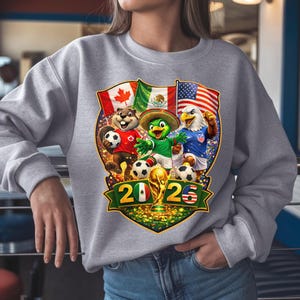 May include: A light gray crewneck sweatshirt featuring a graphic of a beaver, a parrot, and an eagle playing soccer. The design includes the flags of Canada, Mexico, and the USA, along with the text 2026 and a gold trophy centered at the bottom.
