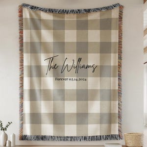 May include: A woven throw blanket featuring a beige and gray buffalo plaid pattern with fringed edges. The center displays the black text "The Williams" in a script font above the text "Forever 02.14.2024" in a simple serif typeface.