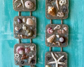 Può includere: Un set di sei decorazioni da parete quadrate a tema nautico. Ogni quadrato è riempito di sabbia e conchiglie, tra cui stelle marine, ricci di mare e vari tipi di conchiglie. I quadrati sono collegati da uno spago e hanno nappe in basso.