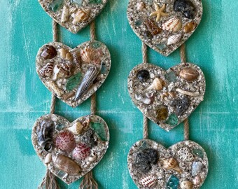 Può includere: Un set di sei ornamenti a forma di cuore realizzati in resina e riempiti di sabbia, conchiglie e vetro di mare. Ogni ornamento ha un fiocco di juta ed è infilato su uno spago.
