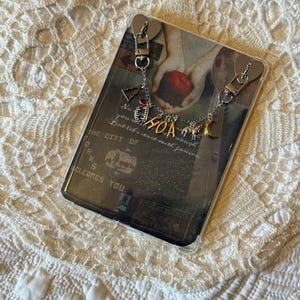 Puede incluir: Una funda transparente para teléfono con un diseño impreso que presenta una pintura de manos sosteniendo una manzana. La funda tiene herrajes plateados y un dije con las letras "HOA", un árbol de Navidad y una luna creciente. El texto "THE CITY OF" es visible.