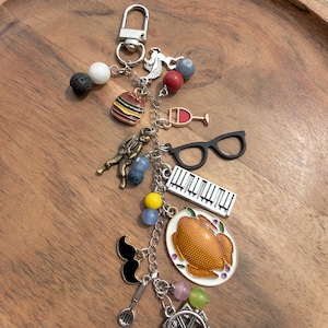 May include: A silver keychain with various charms, including a hamburger, wine glass, eyeglasses, piano keys, a roasted turkey, and a Ferris wheel. The charms are attached to a silver chain with a lobster clasp.